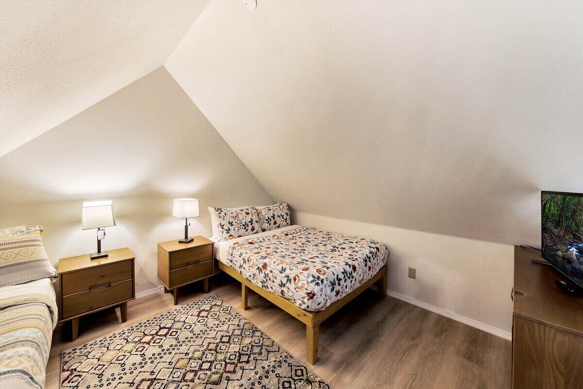 A bedroom with a sloped ceiling, a bed with a floral bedspread, two bedside tables with lamps, a patterned rug, and a television on a wooden cabinet.