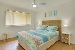 Quarto com cama, ventoinha no teto, janela com persianas, mesa de cabeceira e uma pequena mesa.