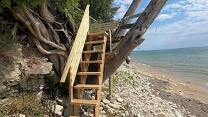 Una escalera de madera que sube por una playa rocosa, con un banco de madera y una estructura de madera apoyada contra un árbol.