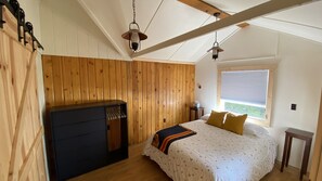 Ett sovrum med träväggar, en säng med ett mönstrat överkast, två sänglampor, en byrå och ett fönster med persienner.