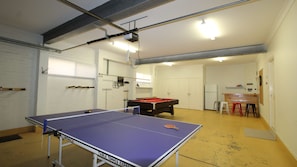A room with a ping pong table, a pool table, and a kitchen area with stools.