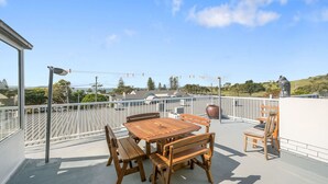 A rooftop terrace with a wooden table and chairs, a metal railing, and a view of the ocean and hills. - Bombora, 3/4 East Street (Crescent Head)