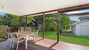 Een overdekt terras met tafel en stoelen, omgeven door een weelderige tuin en een witte garage.