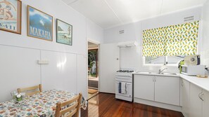 A compact kitchen with white cabinets, a stove, and a microwave. There is a dining table with a colorful tablecloth and a chair. Two framed pictures are hung on the wall. - Herb s Hideaway, 11 Belmore Street (Crescent Head)