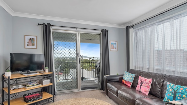 A living room with a television, a sofa with colorful cushions, a bookshelf, and a sliding glass door leading to a balcony.