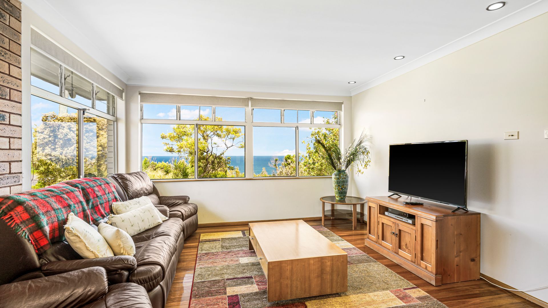 A living room with a leather sofa, a coffee table, a TV on a wooden stand, and a large window with a view of the sea.