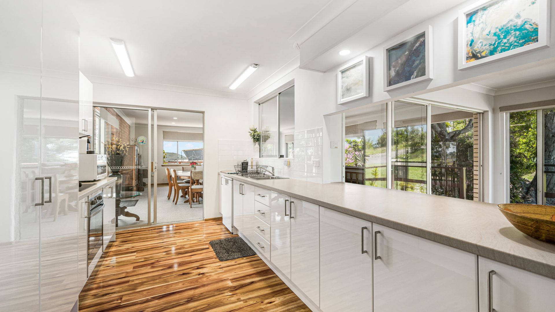 A modern kitchen with a wooden floor, white cabinetry, and large windows offering a view of the outdoors.