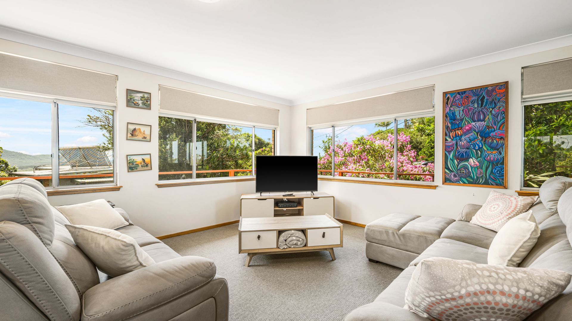 Uma sala de estar moderna com televisão de tela plana, sofás bege, janelas amplas com vista para áreas verdes e obras de arte nas paredes.