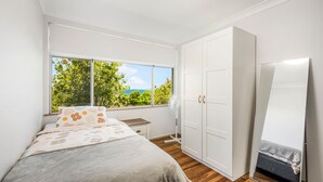 A bedroom with a bed, a nightstand, a wardrobe, a window with a view of greenery and the sea, and a fan. - Beachcomber, 27 Dulconghi Street (Crescent Head)
