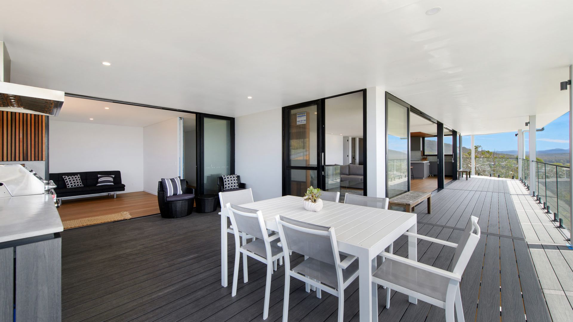 Un espace de restauration extérieur moderne, avec une table et des chaises blanches, une terrasse en bois et une cuisine fermée par des parois de verre.
