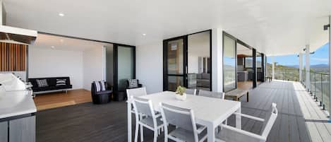 A modern outdoor dining area with a white table and chairs, a wooden deck, and a glass-enclosed kitchen.