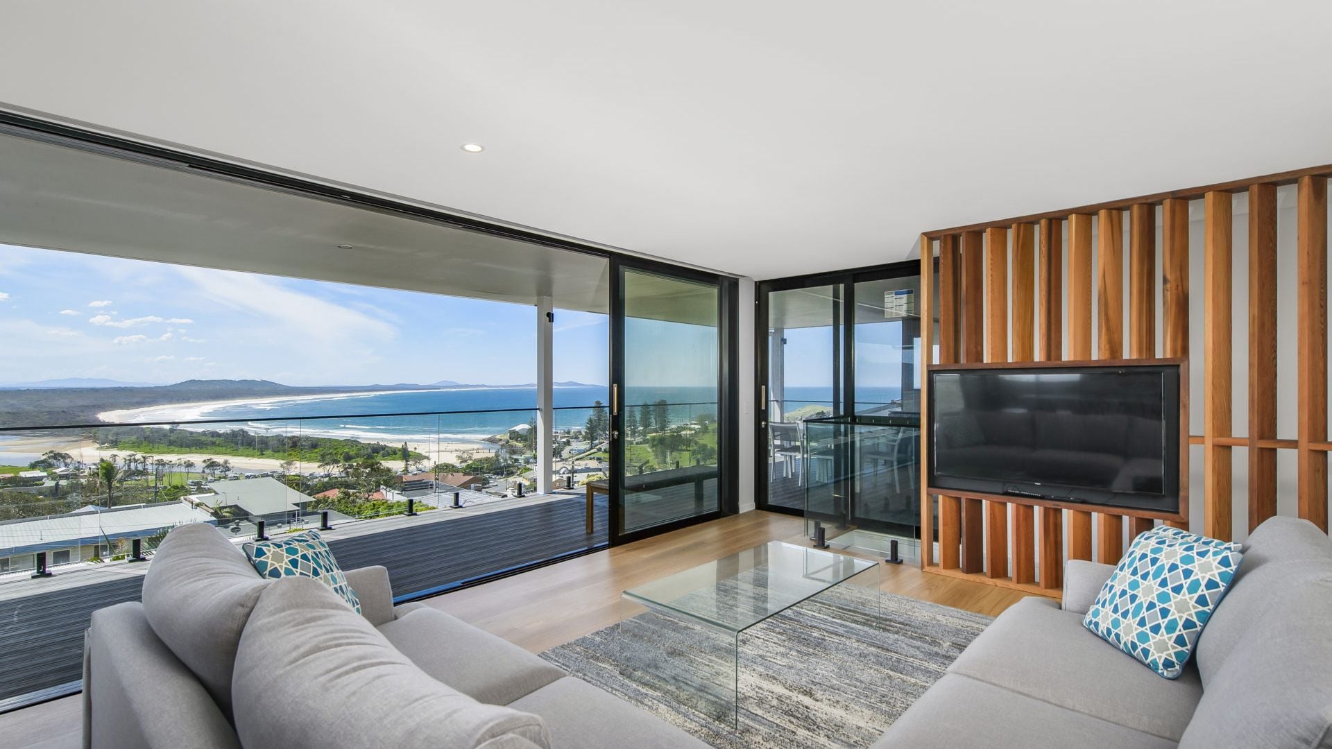 A modern living room with a large glass door leading to a balcony, a flat-screen TV mounted on a wooden panel, and a view of a beach and ocean.