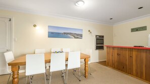 A dining area with a wooden table and white chairs, a painting of a coastal landscape, a wooden counter with a red top, and a wall-mounted air conditioning unit.