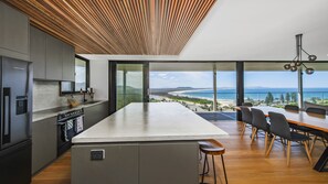 A modern kitchen with a large island, a dining area with a view of the beach, and a wooden ceiling.