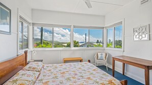 A bedroom with a large bed, a chair, a small table, and multiple windows offering a view of greenery and hills. - Sea Salt, 24 Stewart Street (Crescent Head)