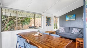 A dining area with a wooden table and blue chairs, a grey sofa with patterned cushions, and a view of a tree and brick wall through large windows. - Sea Salt, 24 Stewart Street (Crescent Head)