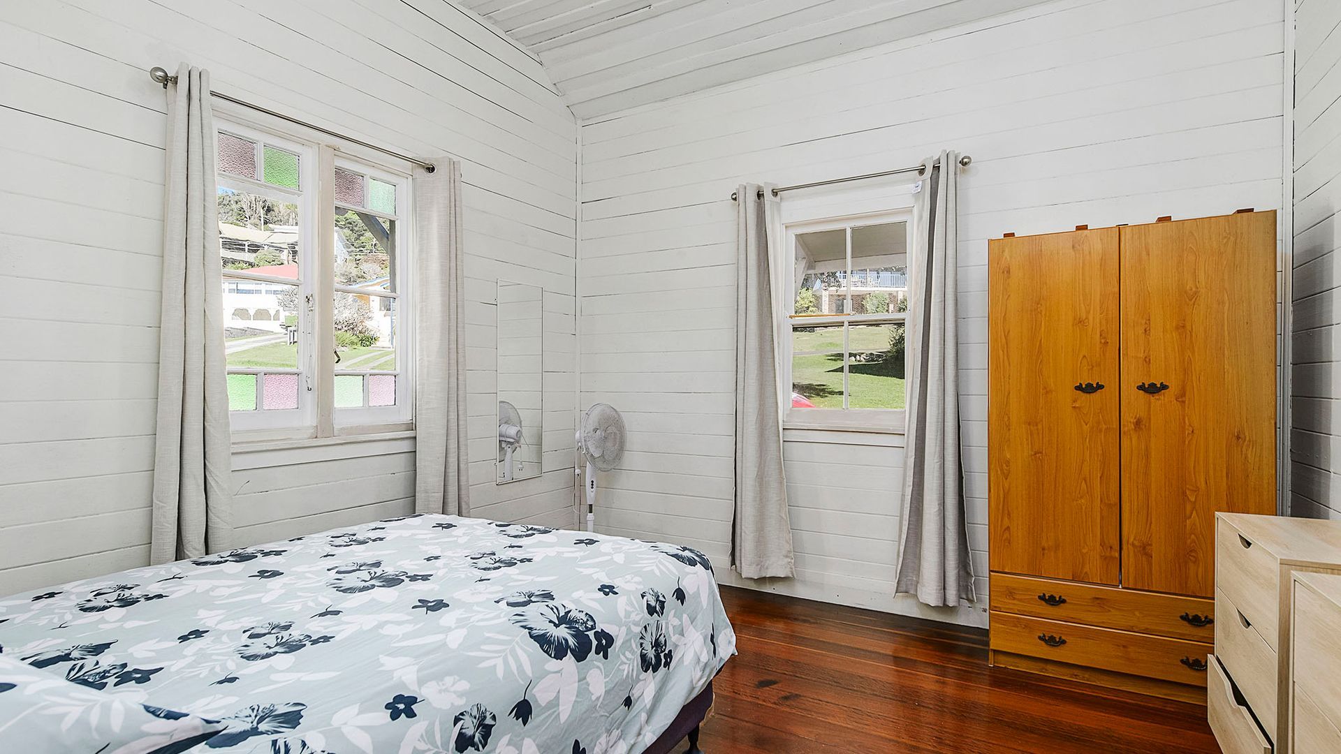 A bedroom with a bed, two windows, a wooden wardrobe, and a fan.