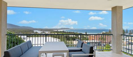 A balcony with a table and two sofas, overlooking a beach and ocean.