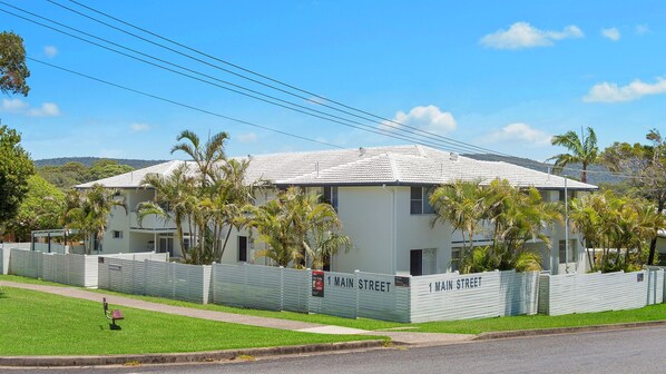 A two-story building with a white exterior and a tiled roof, surrounded by a white fence and palm trees.