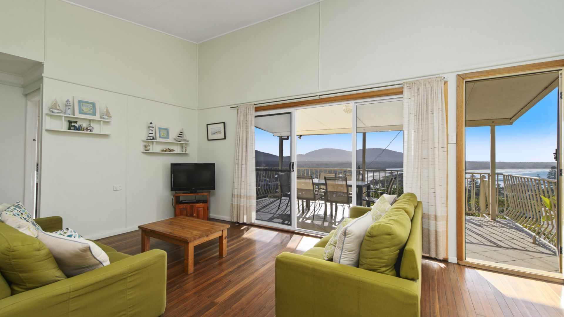 A living room with a wooden floor, green sofa, a flat-screen TV, and a sliding glass door leading to a balcony with a view of mountains.