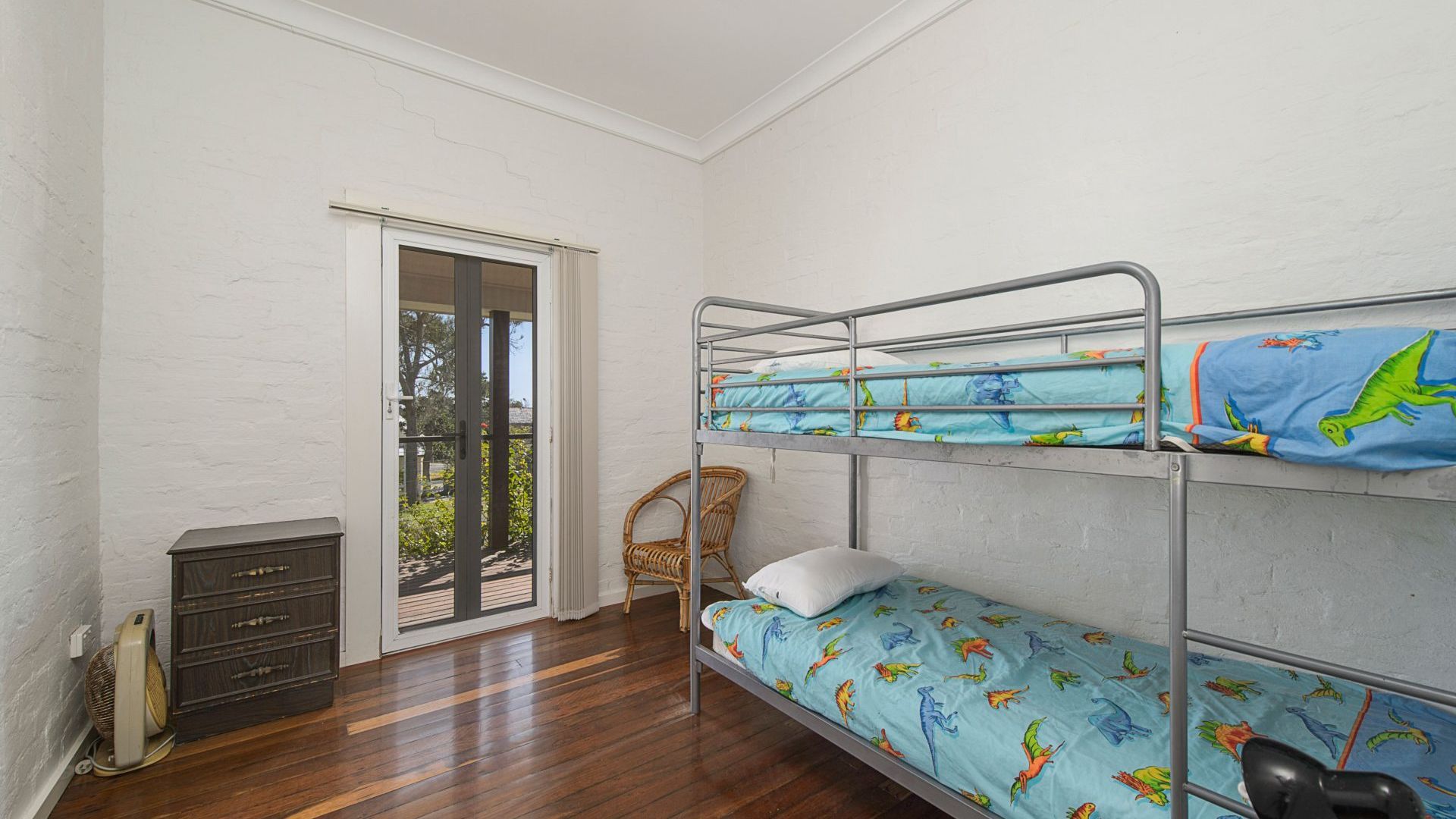Un letto a castello con lenzuola a tema dinosauri, una sedia in vimini, un piccolo comò e una porta scorrevole in vetro che dà su uno spazio all'aperto.