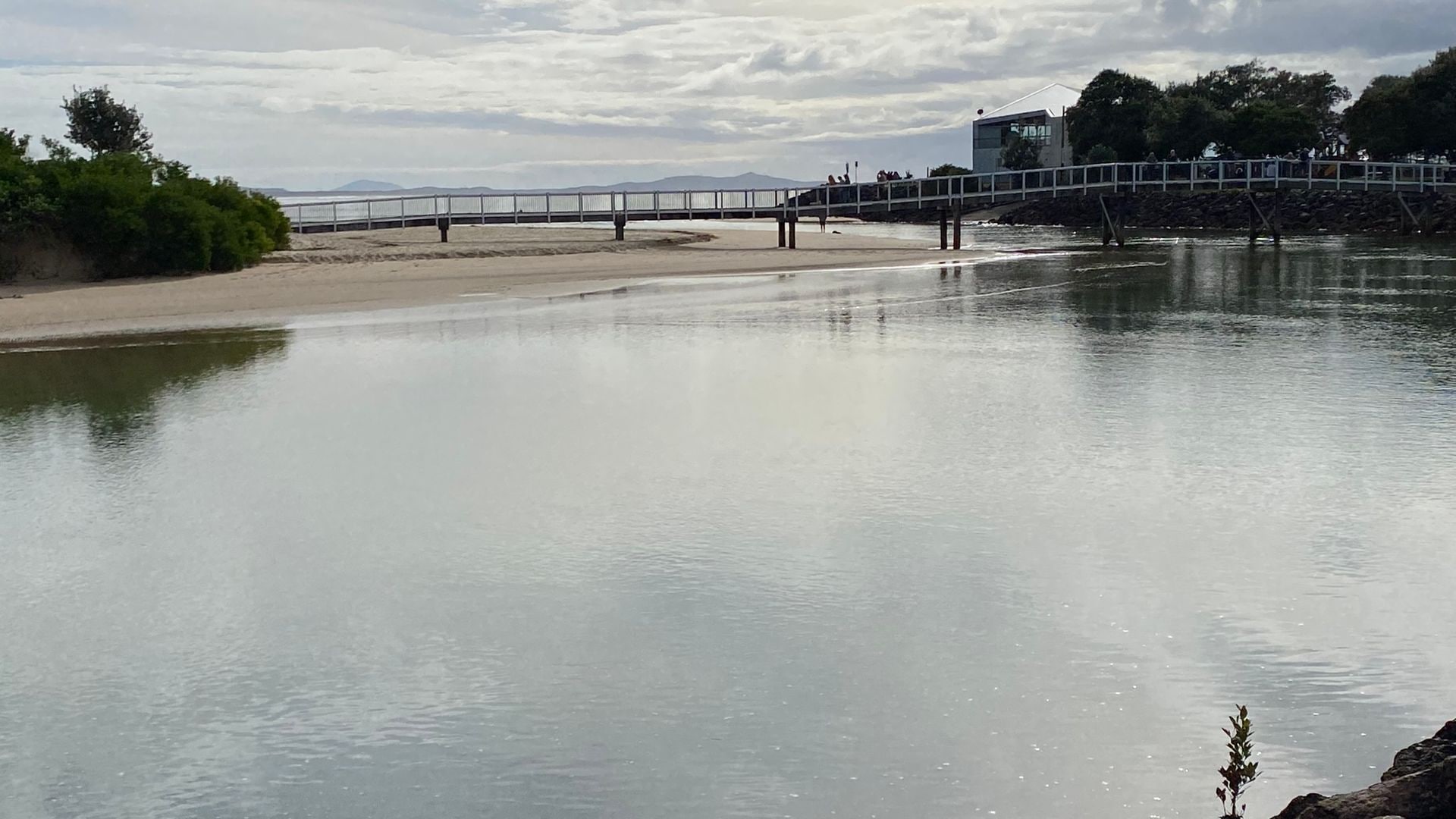 A calm river with a bridge, a sandy beach, and a building in the background.