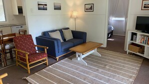 A living room with a sofa, a wooden chair, a coffee table, a lamp, and a television. - Fleur Cottage, 7 Willow Street (Crescent Head)