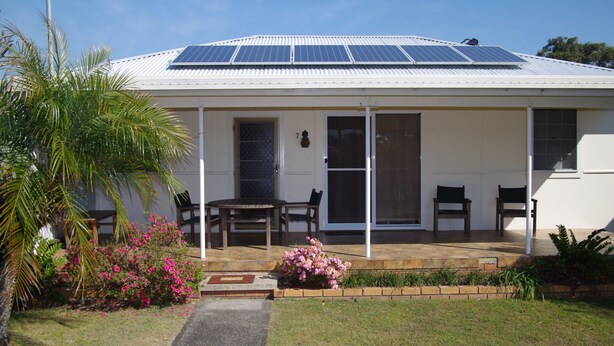 A house with a covered porch, solar panels on the roof, and a small garden with pink flowers. - Fleur Cottage, 7 Willow Street (Crescent Head)