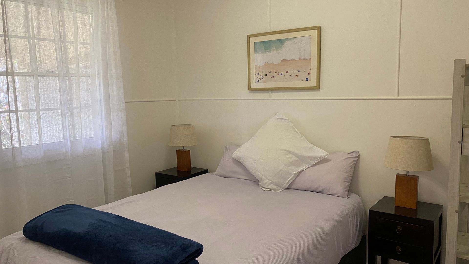 A hotel room with a bed, two bedside tables, a lamp on each table, a window with sheer curtains, and a painting on the wall.