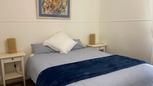 A bedroom with a bed, two bedside tables, a nightstand, a painting on the wall, and a lamp on each table. - Fleur Cottage, 7 Willow Street (Crescent Head)
