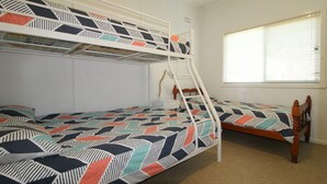Een kamer met stapelbedden, een ladder, een raam met zonwering en een nachtkastje met een lamp.