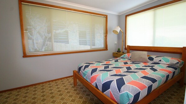 A bedroom with a bed, two windows with blinds, a bedside lamp, and a patterned carpet.