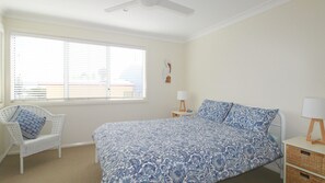 A bedroom with a bed, a chair, a bedside table, a lamp, and a window with blinds. - Beach Club 2, 4 KILLICK LANE (Crescent Head)
