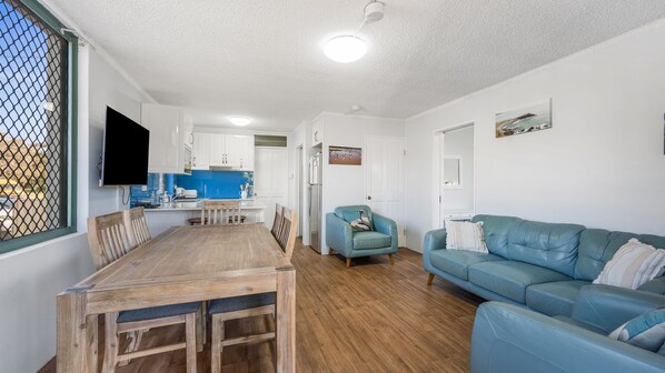 A compact living space with a dining area, a sofa, a television, and a kitchen in the background. - Beachfront 1, 25 Willow Street (Crescent Head)