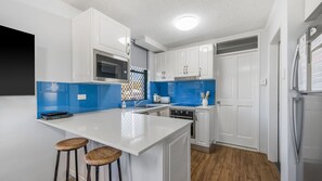 A modern kitchen with white cabinetry, a central island with stools, a microwave, and a refrigerator. - Beachfront 1, 25 Willow Street (Crescent Head)