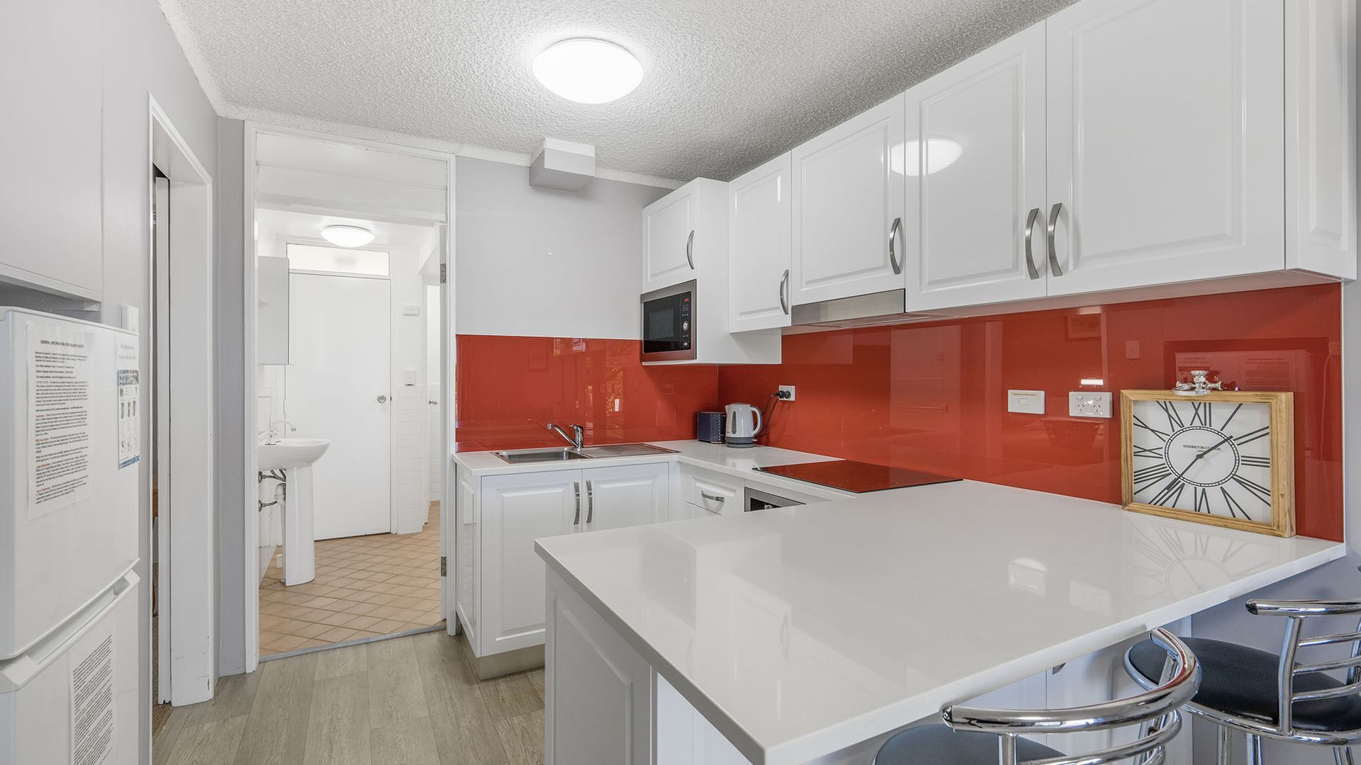A modern kitchen with white cabinets, a red backsplash, and a wooden-framed clock.