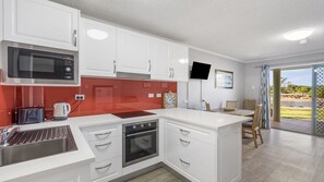 A modern kitchen with white cabinets, a built-in microwave, and an oven. There is a dining area with a table and chairs, and a view of the outdoors through a glass door.