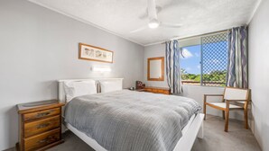 Un dormitorio con cama, escritorio, silla, ventilador de techo, ventana con cortinas e imagen enmarcada en la pared.