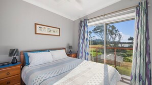 Un dormitorio con cama, mesita de noche, lámpara, cuadro en la pared y un ventanal con cortinas.