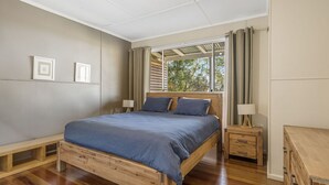A bedroom with a wooden bed, two bedside tables, a window with curtains, and a view of trees. - Beau Soleil, 28 East Street (Crescent Head)