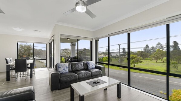 A modern living room with a leather sofa, a dining table, and a large window offering a view of a road and greenery.