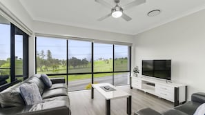 A modern living room with a large window, a flat-screen TV, and a ceiling fan.