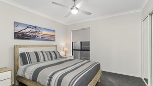 Une chambre à coucher comprenant un lit, une table de chevet, une lampe, un ventilateur de plafond, un tableau et une porte donnant sur une autre pièce.
