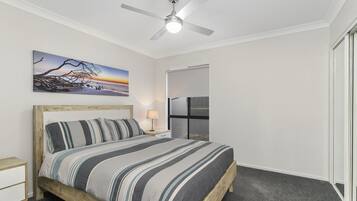 A bedroom with a bed, a nightstand, a lamp, a ceiling fan, a painting, and a door leading to another room.