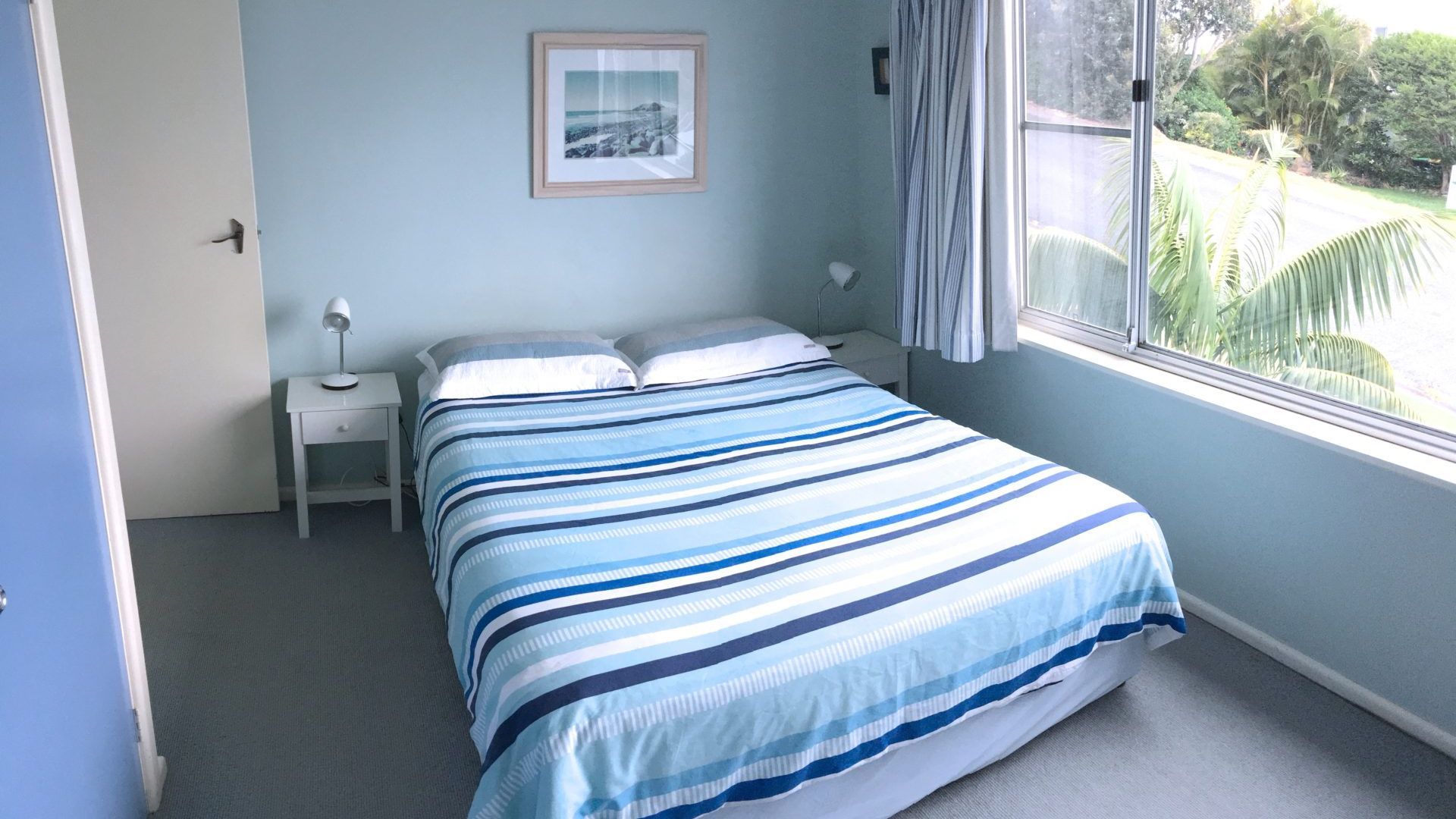Une chambre à coucher avec un lit, une table de chevet, une lampe, un tableau encadré au mur et une fenêtre donnant sur un paysage verdoyant.