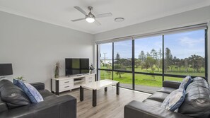 A modern living room with a flat-screen TV, a ceiling fan, and a large window offering a view of a park.