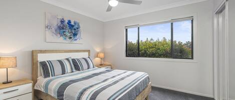 A bedroom with a bed, a ceiling fan, a window with a view of greenery, and a painting on the wall.
