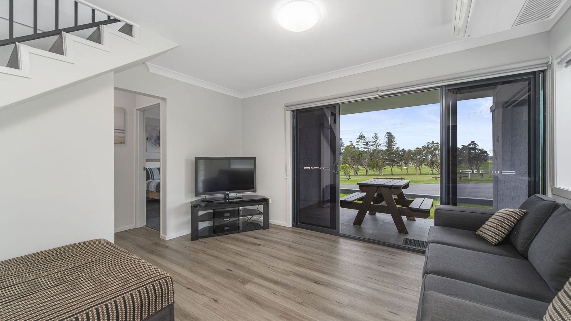 A modern living room with a flat-screen TV, a sofa, a wooden bench, and a patio area with a table and chairs.