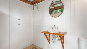 A bathroom with white tiled walls, a wooden vanity with a sink, a mirror, and a shower area. - The Tree House, 6 Gowing Street (Crescent Head)