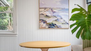 A room with a round table, a framed painting on the wall, and a window with a view of greenery.